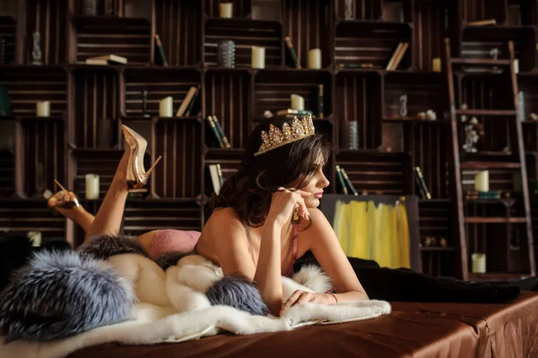 lateral view of a sexy woman laying on a white fur coat with gray collar, in pink underwear with a crown on her head, near bookshelves with books