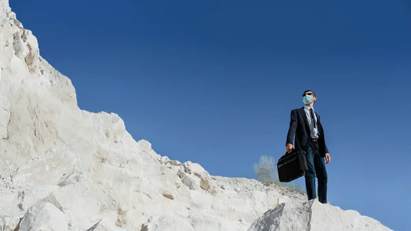 Young man businessman with a briefcase and face mask is climbing in the mountain. Concept of coronavirus COVID 19 pandemic and economic crisis.