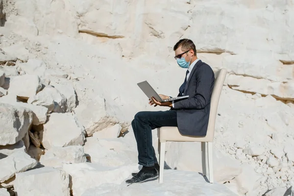 Young man businessman with a face mask is sitting on a chair and working on a notebook computer in the mountain in Coronavirus Covid 19 Pandemic. Concept of online work in coronavirus COVID 19 pandemic. Work from everywhere