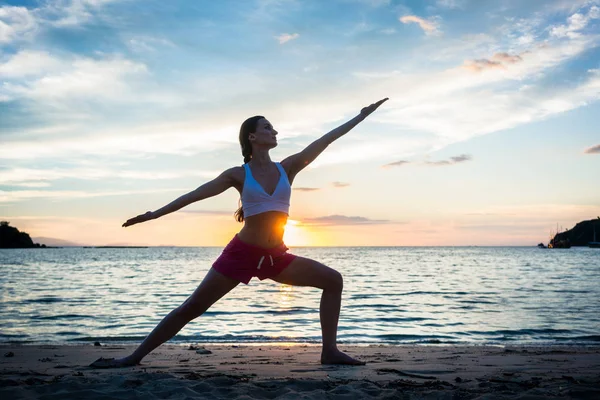 Silhueta de uma mulher apta praticando a pose de ioga guerreira contra o pôr do sol — Fotografia de Stock