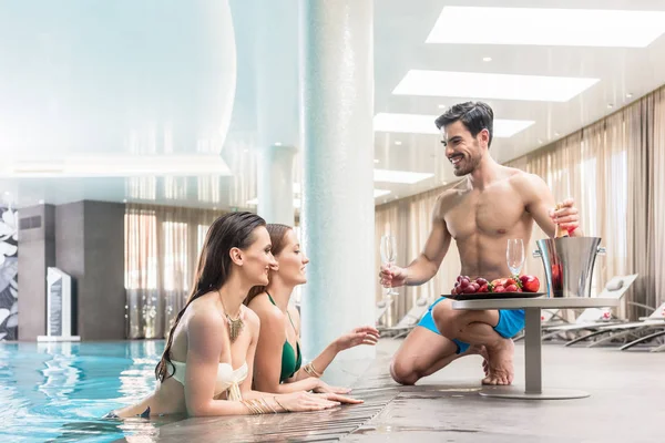 Young man serving with champagne two women at the swimming pool