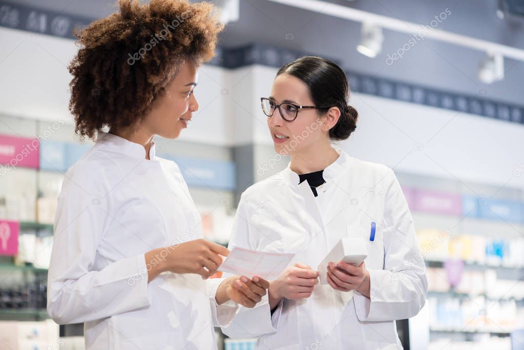 Two female pharmacists reading the information from the pack of a new product while comparing two different medicines regarding indications and side effects 