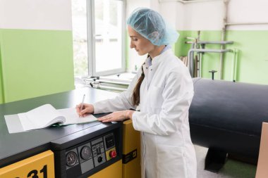 Ekipman ve duran yakınındaki kompresör makine bir modern kozmetik fabrikasında üretim hakkında gözlemler bir rapor için yazılı bir kadın mühendisi yan görünüm