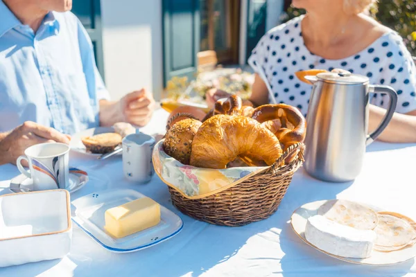 Двоє людей старшого віку снідають з хлібним кошиком на столі в літньому саду — стокове фото