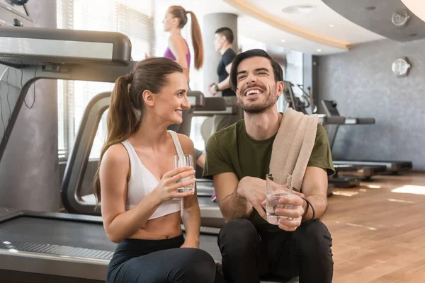 Alegre joven y joven bebiendo agua corriente durante el descanso en el gimnasio — Foto de Stock