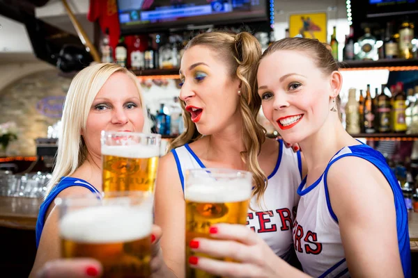 Девушки из группы поддержки смотрят игру в спортивном баре, пьют пиво — стоковое фото