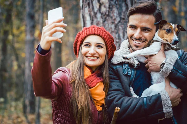 Mulher e homem com seu cão na caminhada de outono tomando uma selfie de telefone — Fotografia de Stock