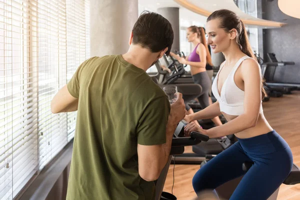 Personal trainer setting the timer of the stationary bicycle for his client — Stock Photo, Image