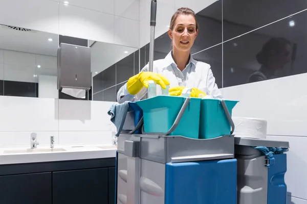 Уборщица или уборщица со своими рабочими инструментами смотрит в камеру в туалете — стоковое фото