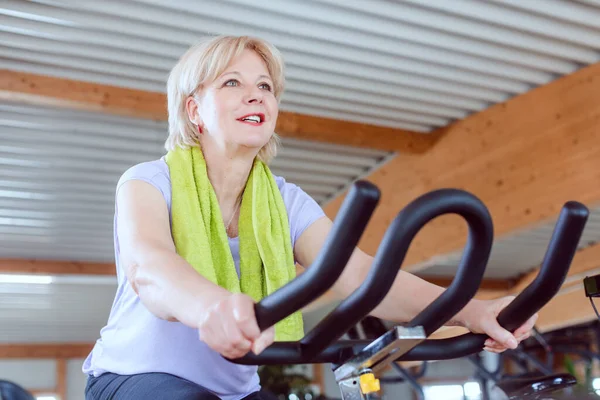 Mulher sênior em uma bicicleta de exercício no ginásio — Fotografia de Stock