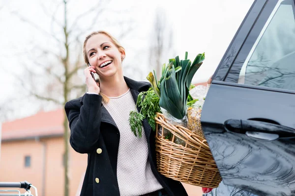 Wanita menggunakan telepon sambil menyimpan bahan makanan di bagasi mobil — Stok Foto