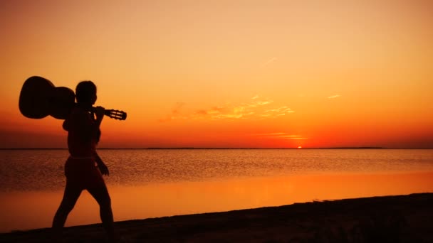 Ung flicka promenerar med gitarr på stranden vid solnedgången — Stockvideo