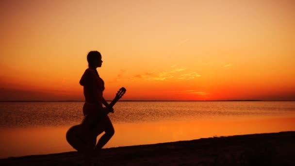 Młoda dziewczyna spaceru z gitarą na plaży o zachodzie słońca — Wideo stockowe