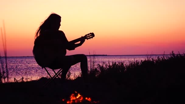 Jovem tocando guitarra na praia com fogueira ao pôr do sol — Vídeo de Stock