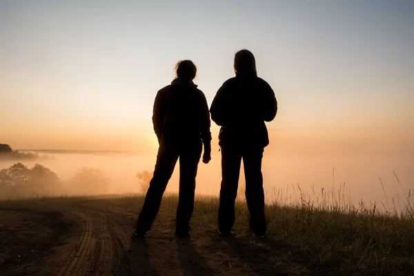 Silhouette of two peple looking at sunrise on foggy valley — Stock Photo, Image