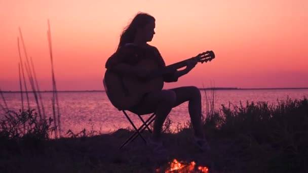 Молодая девушка играет на гитаре на пляже с костром на закате — стоковое видео