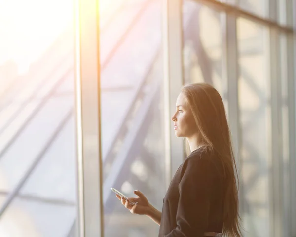 Succesvol zakenvrouw met een smartphone, staande voor een venster in een modern kantoor. — Stockfoto