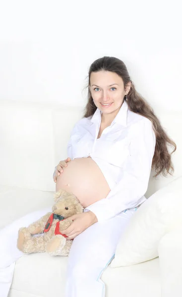 Sorrindo mulher grávida sentado no sofá com ursinho de pelúcia — Fotografia de Stock