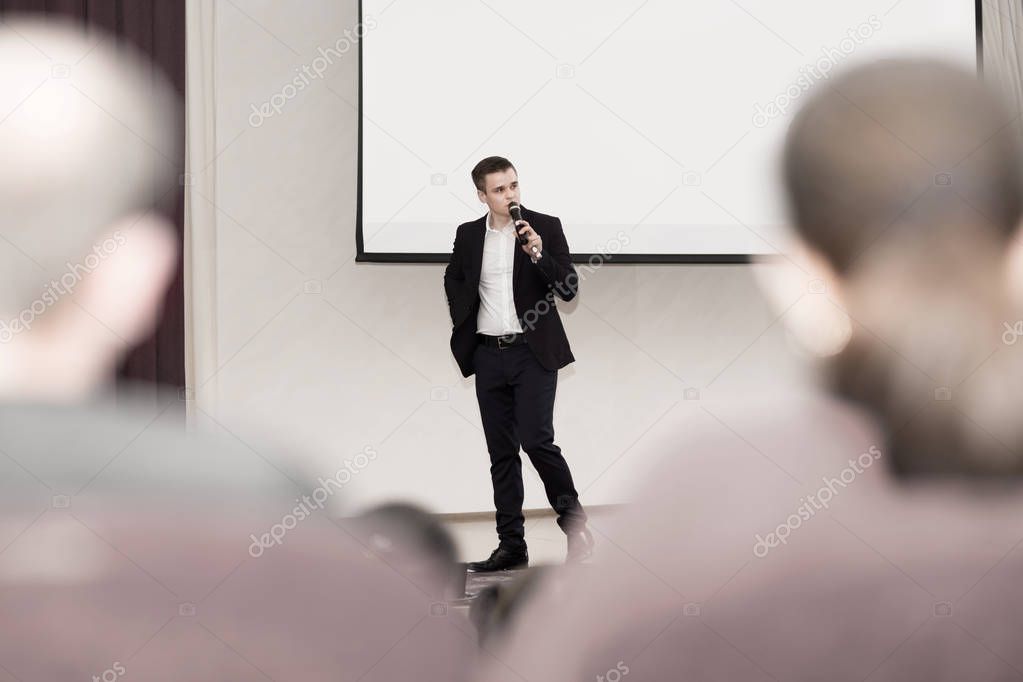 speaker conducts the business of the conference standing in front of a large white screen on the stage in the conference room