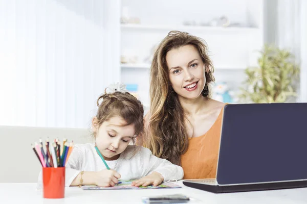 Joven madre - mujer de negocios que trabaja en el ordenador portátil y su hija de cinco años dibuja con lápices de colores —  Fotos de Stock