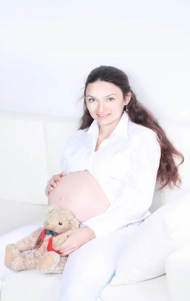 Donna incinta sorridente seduta sul divano con orsacchiotto — Foto Stock