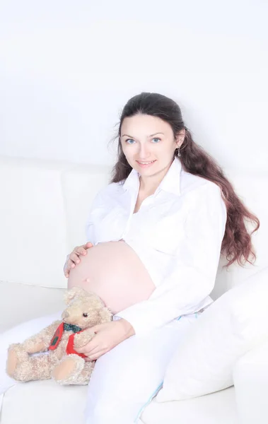 Sorrindo mulher grávida sentado no sofá com ursinho de pelúcia — Fotografia de Stock