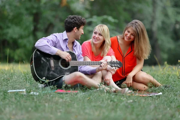 three students with a guitar sitting on the grass in the city Park