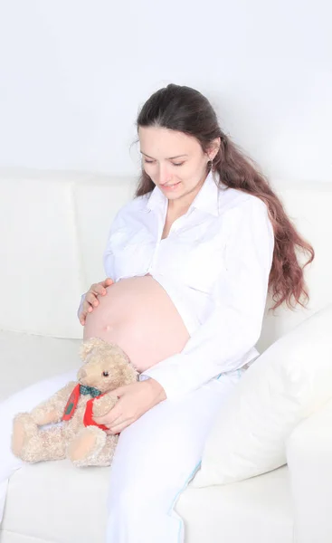 Sorrindo mulher grávida sentado no sofá com ursinho de pelúcia — Fotografia de Stock