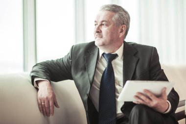 kanepede oturan ve ofis pencereden dışarı bakarak dijital tablet ile deneyimli iş adamı