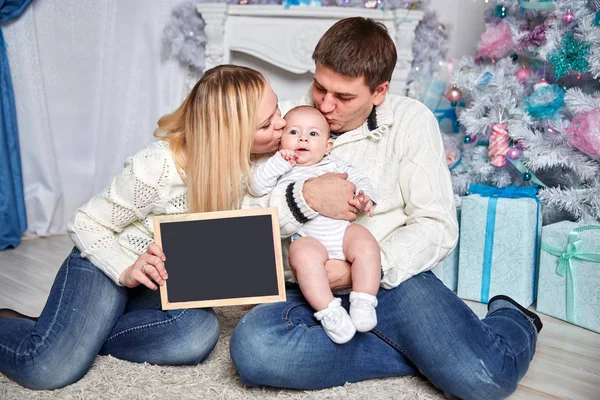 Familia feliz sosteniendo una tarjeta de Navidad en blanco, sentado en la alfombra en una acogedora sala de estar — Foto de Stock