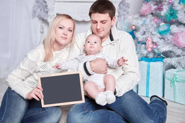 Happy family holding a blank Christmas card, sitting on the carpet in a cozy living room — Stock Photo, Image