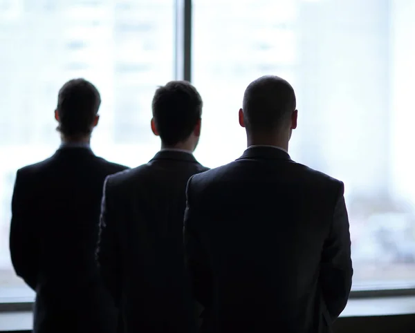 Widok z trzech biznesmenów z tyłu jak oni patrzą na duże okna z widokiem na miasto — Zdjęcie stockowe