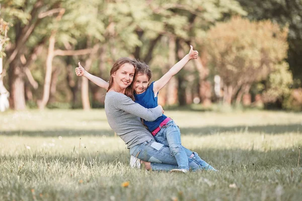 Feliz mamá abrazando a su pequeña hija sentada en el césped el día de verano —  Fotos de Stock