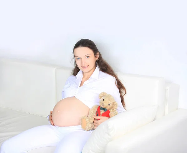 Sorrindo mulher grávida sentado no sofá com ursinho de pelúcia — Fotografia de Stock