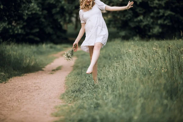 Glückliche schwangere Frau mit einem Blumenstrauß, die barfuß im Gras spaziert — Stockfoto