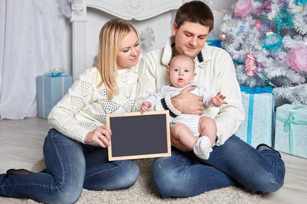 Família feliz segurando um cartão de Natal em branco, sentado no tapete em uma acolhedora sala de estar — Fotografia de Stock