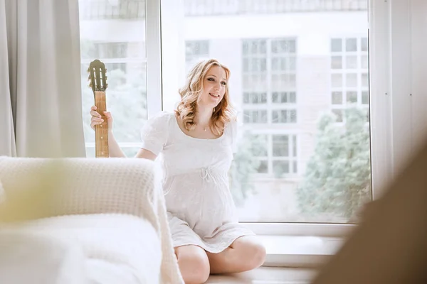 Jeune femme enceinte avec une guitare assise près de la fenêtre dans le salon — Photo