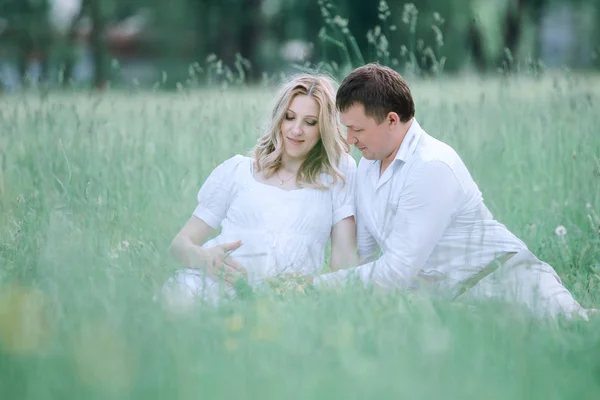 Krásná těhotná žena se svým manželem, sedí na trávníku v letním dni. — Stock fotografie