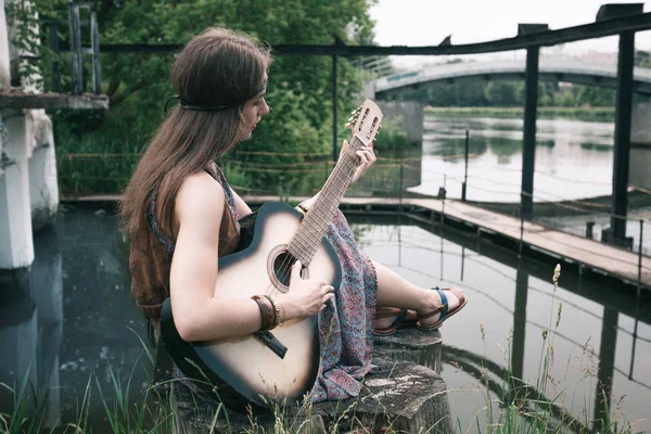 Jovem hippie mulher tocando guitarra sentado perto da lagoa — Fotografia de Stock