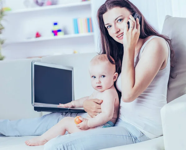 Fiatal anya éves kisgyermeket tartott a karját, beszél a mobiltelefonján, és működő-ra laptop — Stock Fotó