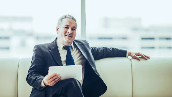 Experienced businessman looking at the screen of a digital tablet sitting on sofa in the office — Stock Photo, Image