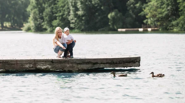 Mamá con su pequeño hijo alimentando patos salvajes en un paseo cerca del lago . —  Fotos de Stock