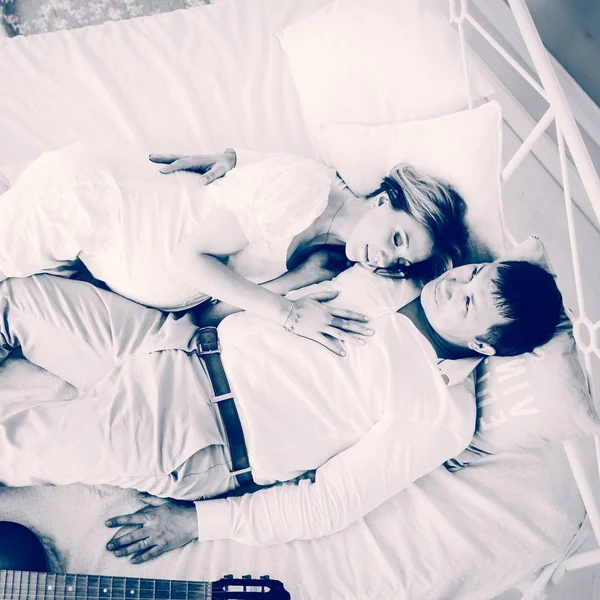 Kochający mąż z gitarą i jego ciężarna żona w łóżku w piękne wnętrza — Zdjęcie stockowe