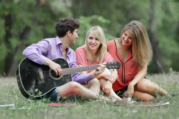 three students with a guitar sitting on the grass in the city Park