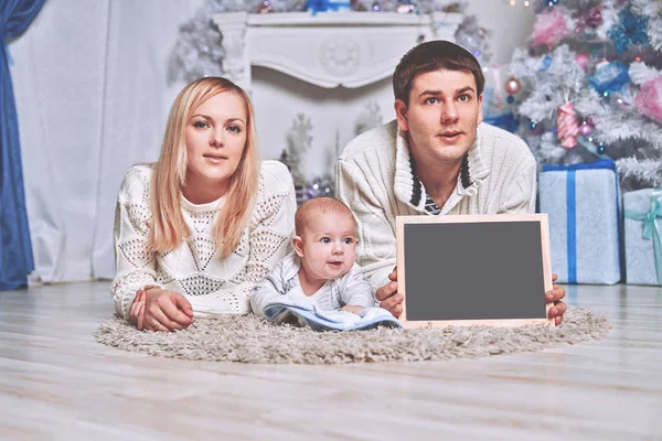 Familia feliz sosteniendo una tarjeta de Navidad en blanco, acostado en la alfombra en una acogedora sala de estar — Foto de Stock