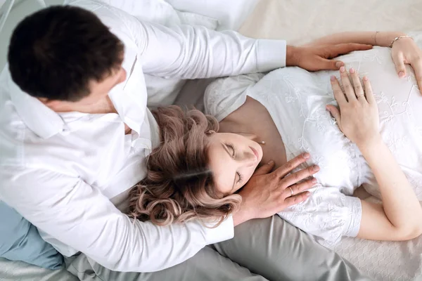 Mann umarmt seine schwangere Frau, während er auf dem Bett sitzt. — Stockfoto