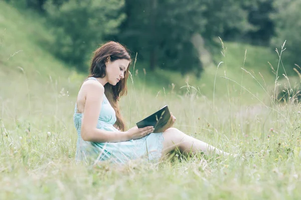 Ładna młoda kobieta z książką siedzącą na zielonej trawie — Zdjęcie stockowe