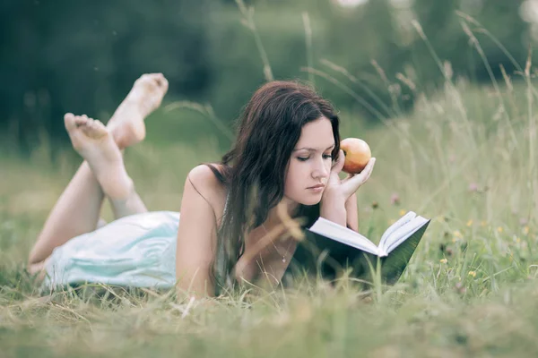 Chica atenta con manzana acostada en la hierba verde y leyendo un libro . —  Fotos de Stock