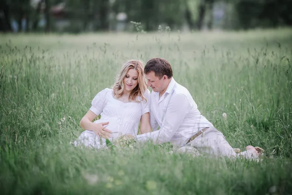 Vacker gravid kvinna med maken sitter på gräsmatta i sommardag. — Stockfoto