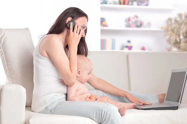 Fiatal anya játszik beszél egy mobiltelefon és gépelés egy laptop — Stock Fotó